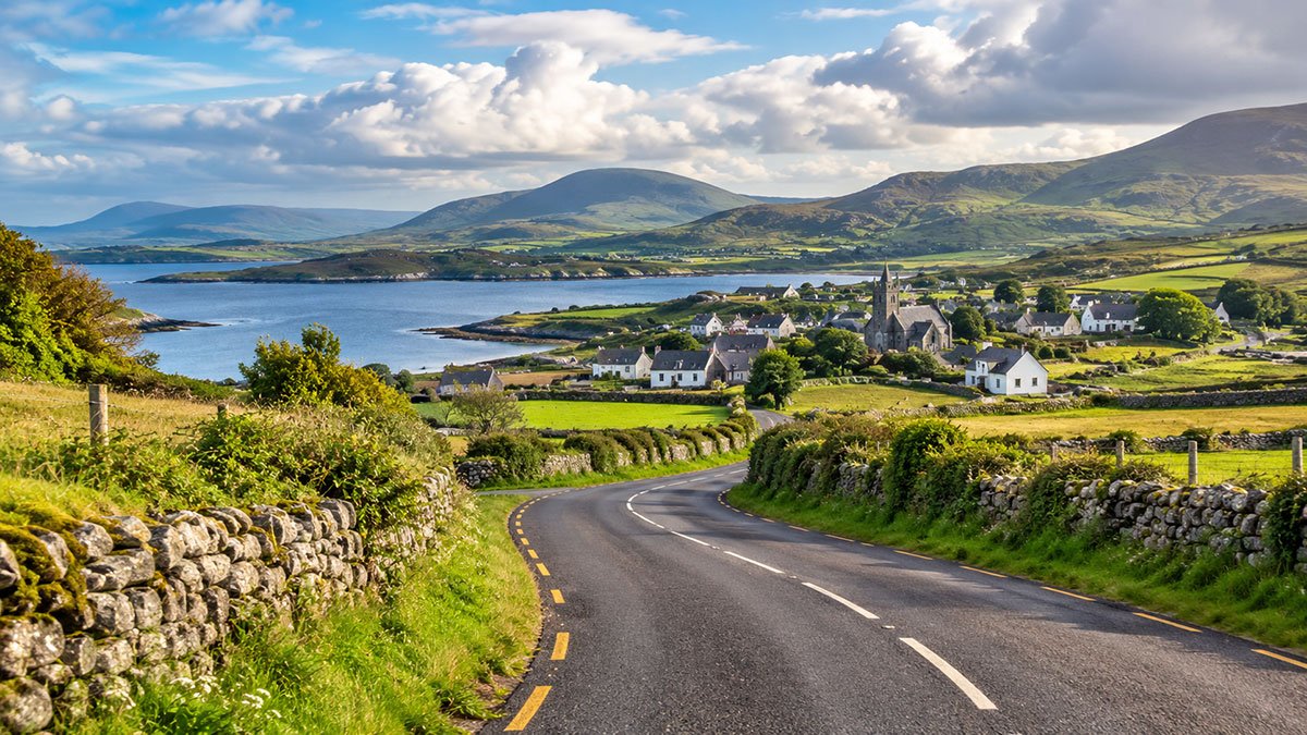Winding country road through Ireland’s green hills, bordered by stone walls and leading to a picturesque coastal village.