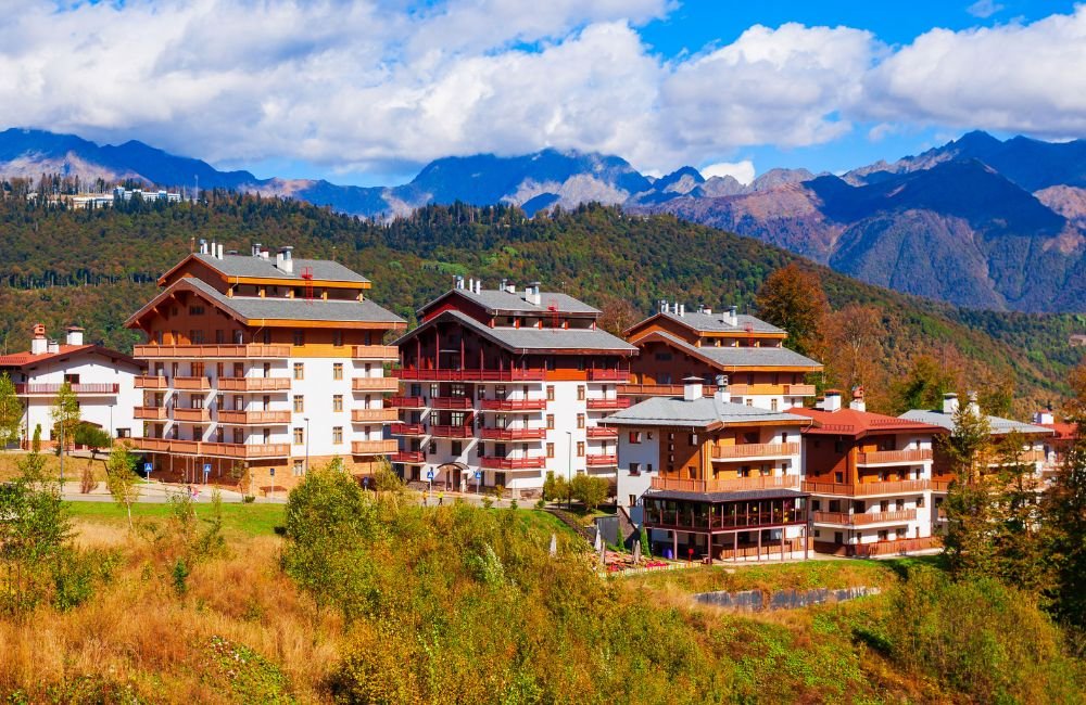 Alpine-style resort buildings at Rosa Khutor near Sochi, Russia, surrounded by forested hills and the Caucasus Mountains.