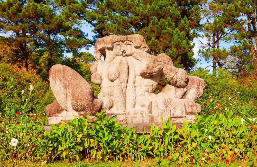 Decorative stone sculpture surrounded by colourful flowers, green plants and tall trees in Sochi Arboretum Park, Russia.