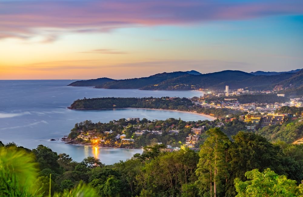 Panoramic sunset view from Karon Viewpoint in Phuket, Thailand, showing curved coastal bays, tropical green hills and the Andaman Sea.