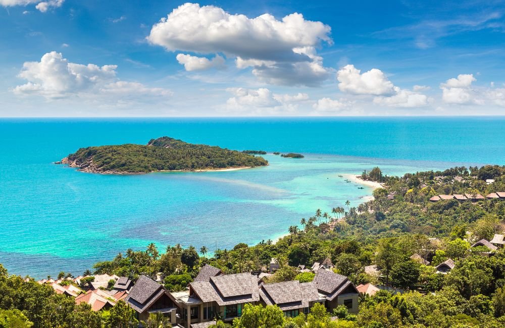 Panoramic coastal view in Koh Samui, Thailand, showing turquoise sea, a small tropical island, palm trees and hillside resort villas.
