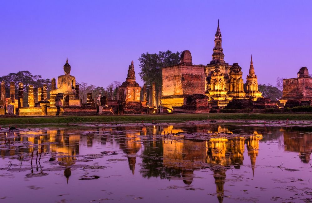 Illuminated ancient temple ruins, Buddha statue and historic chedis reflected in a pond at Sukhothai Historical Park, Thailand, at dusk.