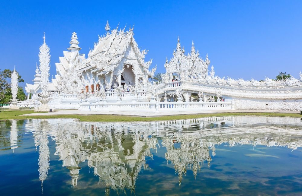 Wat Rong Khun White Temple in Chiang Rai, Thailand, featuring intricate white carvings, ornate spires and reflections in a tranquil pond.